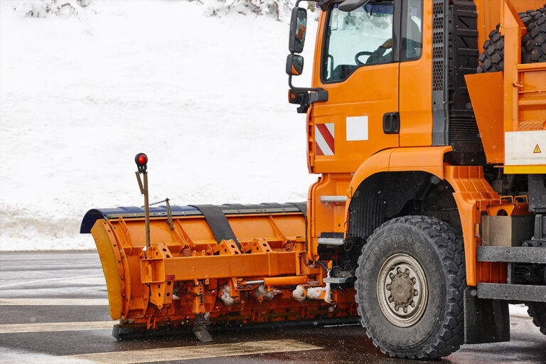 Ein orangefarbener Schneepflug steht auf einer nassen Straße und ist mit einer großen Schaufel zum Schneeräumen ausgestattet. Im Hintergrund ist Schnee auf dem Boden und am Straßenrand zu sehen. Die Gärtner im Allgäu