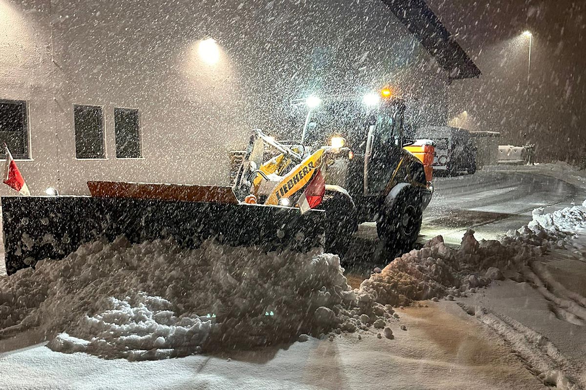 Ein Schneepflug räumt nachts schweren Schnee von einer Straße vor einem Gebäude. Es schneit heftig, und die Gegend wird von hellen Lichtern erhellt, die eine winterliche Szene schaffen. Die Gärtner im Allgäu