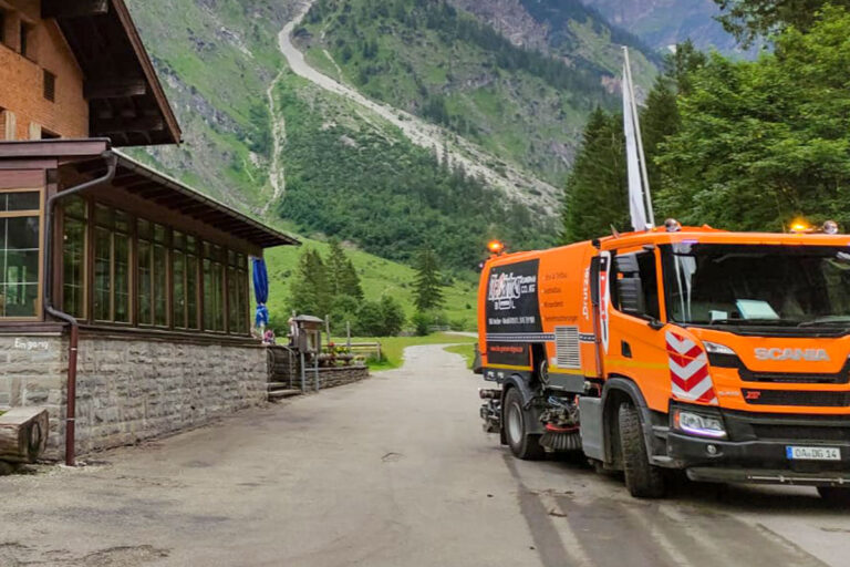 Ein orangefarbener Scania-Straßenreinigungswagen parkt auf einer gepflasterten Straße neben einem Steingebäude, während im Hintergrund grüne Berge und Bäume zu sehen sind. Die Gärtner im Allgäu