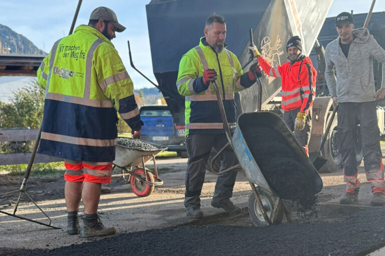 Vier Arbeiter in Sicherheitskleidung verteilen mit Harken und Schubkarren in der Nähe eines Lastwagens Asphalt auf dem Boden, während im Hintergrund an einem sonnigen Tag Bäume und Berge zu sehen sind. Die Gärtner im Allgäu