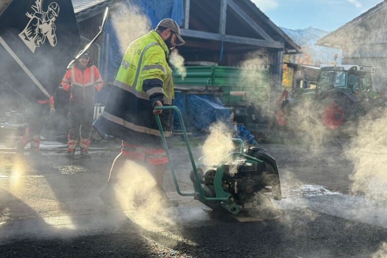 Ein Bauarbeiter in einer gelben Sicherheitsjacke bedient eine Rüttelplatte, umgeben von Dampf oder Staub, während zwei andere Arbeiter und ein Traktor an einem sonnigen Tag im Hintergrund stehen. Die Gärtner im Allgäu