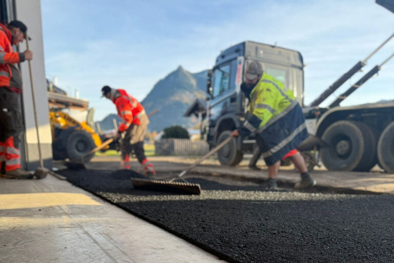 Drei Arbeiter in Schutzkleidung verteilen mit Harken Asphalt auf einer Straße. In der Nähe sind Baufahrzeuge zu sehen, und im Hintergrund ist bei klarem Himmel ein Berg zu erkennen. Die Gärtner im Allgäu