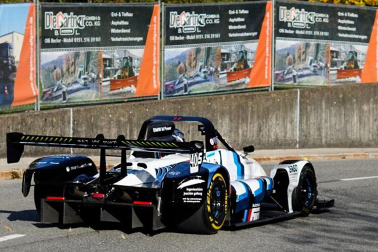 Ein schwarz-weißer Rennwagen mit blauen Akzenten und der Nummer 405 auf der Seite ist auf einer Straße in der Nähe einer Betonmauer mit Werbung im Hintergrund geparkt. Die Gärtner im Allgäu