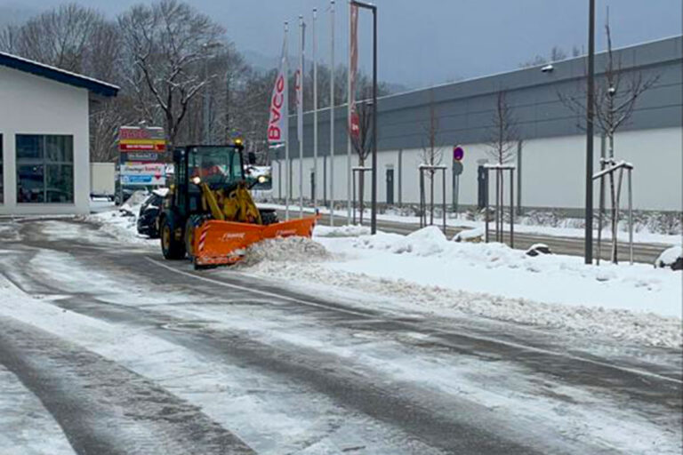 Ein Schneepflug räumt an einem Wintertag Schnee von einer Straße in der Nähe von Geschäftsgebäuden und geparkten Autos; im Hintergrund sind Bäume und Fahnen zu sehen. Die Gärtner im Allgäu