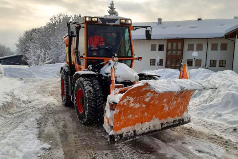 Ein Schneepflug mit orangefarbenem Schild räumt im Winter eine verschneite Straße vor einem Gebäude. Schneebänke säumen die Seiten, und im Hintergrund sind mit Schnee bedeckte Bäume zu sehen. Die Gärtner im Allgäu